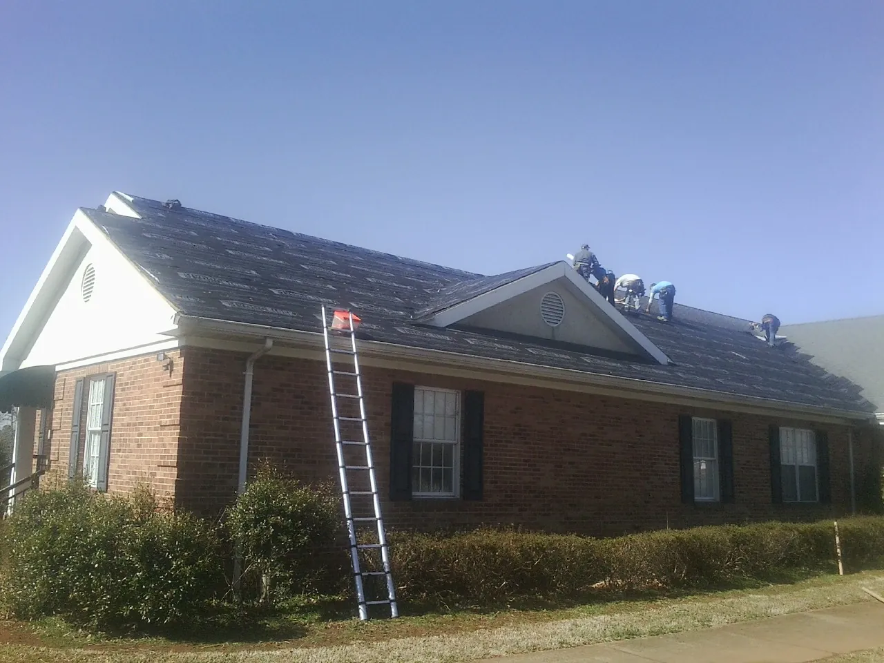Crew tearing shingles off a brick ranch roof with a ladder set — Emergency Roof Repair work by our crew in Gwynn