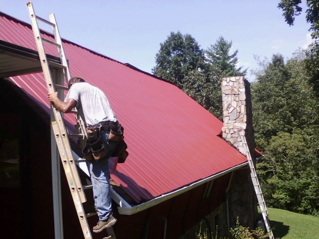 Roofer on a ladder beside a red metal roof and stone chimney — Soffit & Fascia Repair by Roofing Brothers in Gwynn